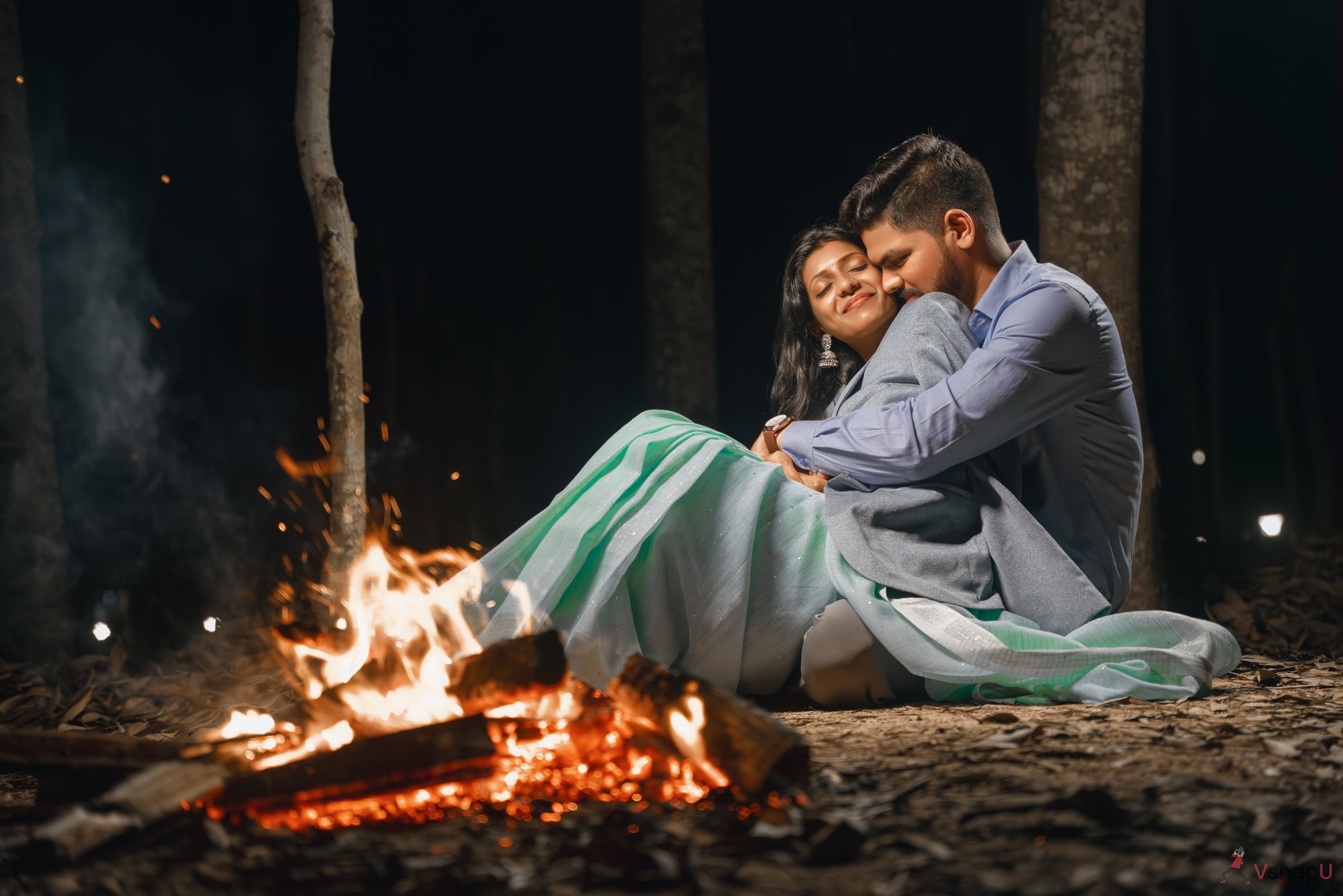 Romantic scene of a couple in traditional attire hugging by a fire.